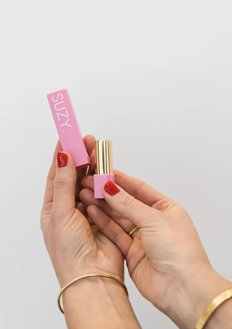 SUZY Whipped Matte Lipstick packaging featuring a soft pink case with white SUZY branding and gold lipstick tube, held in hands against a neutral background.