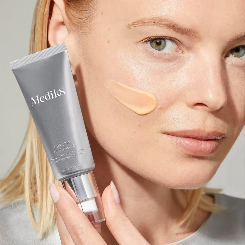 Woman holding Medik8 Crystal Retinal 6 Night Serum 30ml beside her face, with a peach-coloured retinal cream swatch on her cheek.