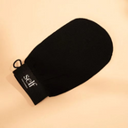 Black SELF Tan by O Cosmedics body exfoliating glove displayed on a beige background, showing its textured fabric.