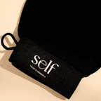Close-up of the SELF Tan by O Cosmedics Exfoliating Glove showing its black fabric, stitching and branded label