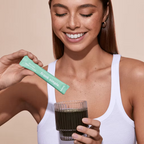Woman holding a Frank Simple Greens Vital Daily Nutrients Mango & Papaya single-serve sachet above a glass of green superfood drink.