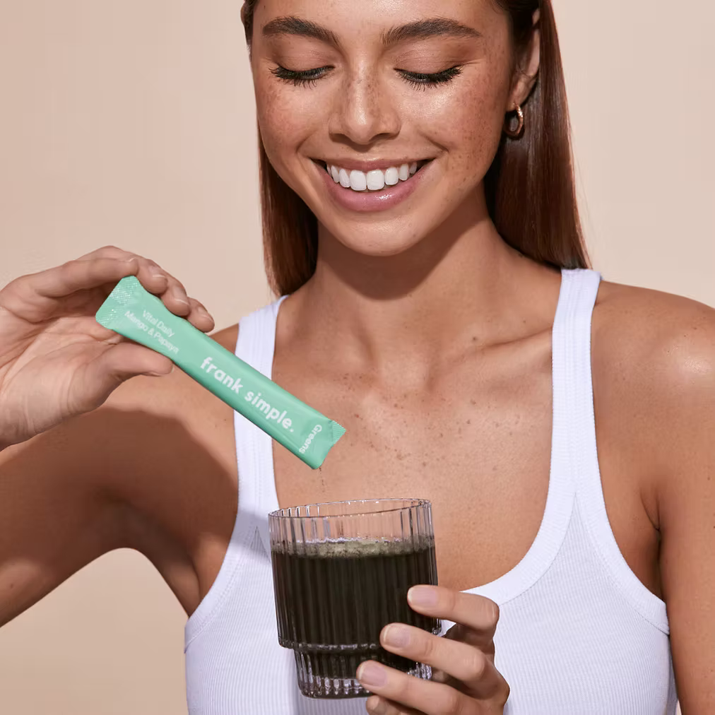 Woman holding a Frank Simple Greens Vital Daily Nutrients Mango & Papaya single-serve sachet above a glass of green superfood drink.