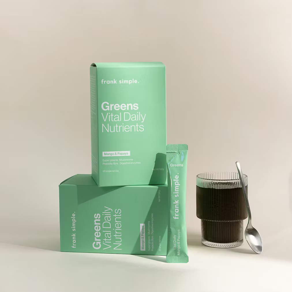 Frank Simple Greens Vital Daily Nutrients Mango & Papaya product boxes and single-serve sachet displayed beside a glass of prepared greens drink with a spoon.