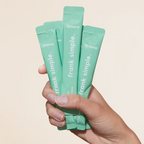 Hand holding Frank Simple Greens Vital Daily Nutrients Mango & Papaya single-serve green superfood powder sachets against a neutral beige background.