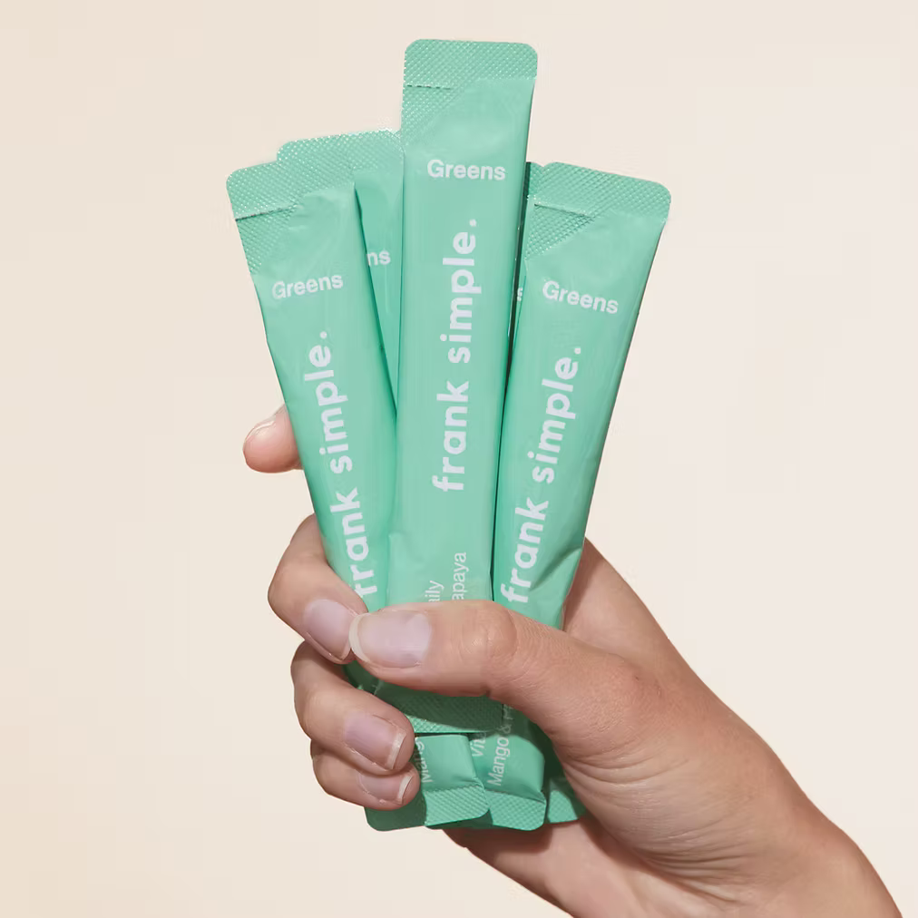 Hand holding Frank Simple Greens Vital Daily Nutrients Mango & Papaya single-serve green superfood powder sachets against a neutral beige background.