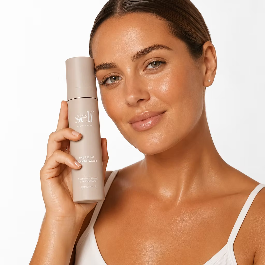 Woman holding SELF Tan by O Cosmedics Hydrating Face Tan Water beside her face, showing a radiant complexion