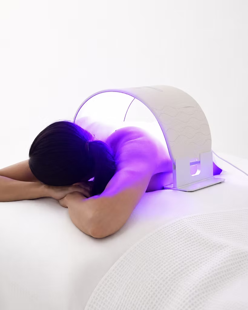 Woman lying face down with the O Cosmedics O LED Light Therapy Device positioned over her upper back and shoulders, demonstrating LED body light therapy.